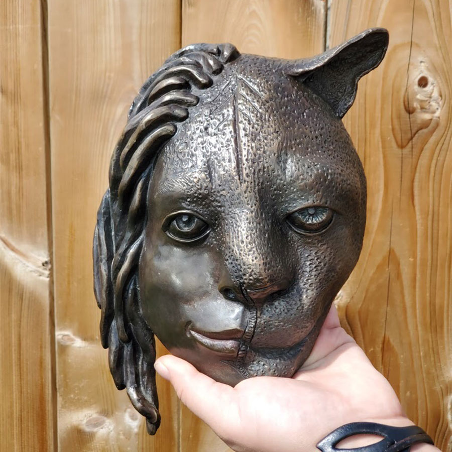 Bronze casted mask that is half human and half cougar. The human half is on the left, has dreadlocks, and is smiling.