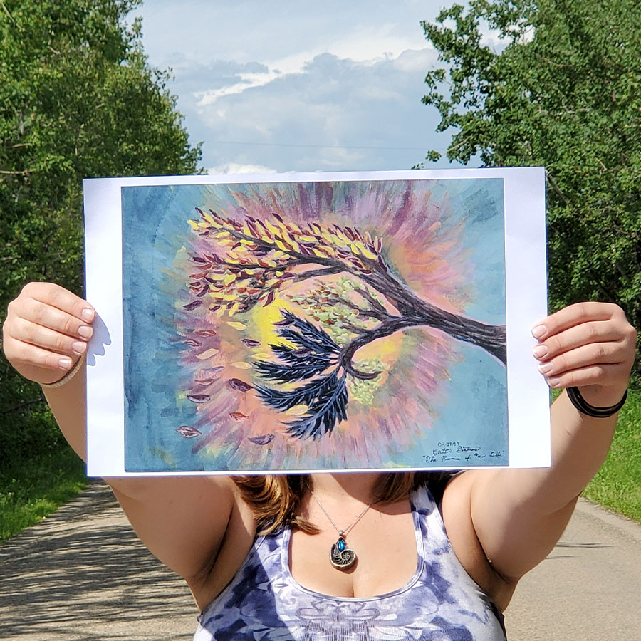 Image of the artist holding up a print of her painting. It has a branch with green leaves, but one area is turning fall colours. The sun is shining through the branch and leaves. There's a branch full of raven feathers.